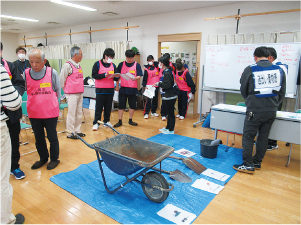 一輪車やスコップなどの防災用品と訓練をしてる生徒と地元の住民たちの写真